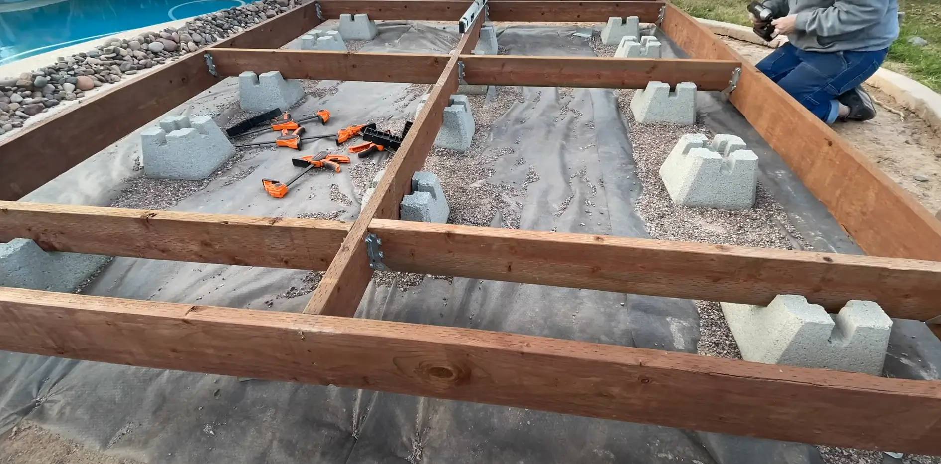 Construction of a floating deck foundation with wooden beams secured on concrete blocks, surrounded by gravel and tools. Build your dream outdoor space with a sturdy floating deck setup!