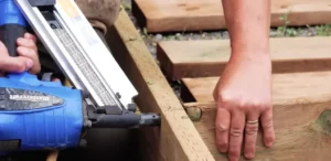 Close-up of a person using a nail gun to construct a wooden deck frame. Ready to build your deck? Contact us for expert decking services!