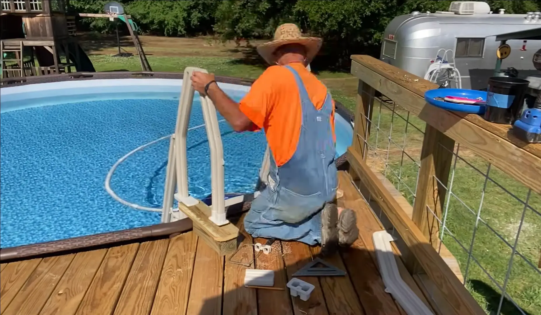 Man in overalls installing a pool ladder onto a wooden deck by an above-ground pool. Secure your pool area safely—hire a professional contractor for expert installation.